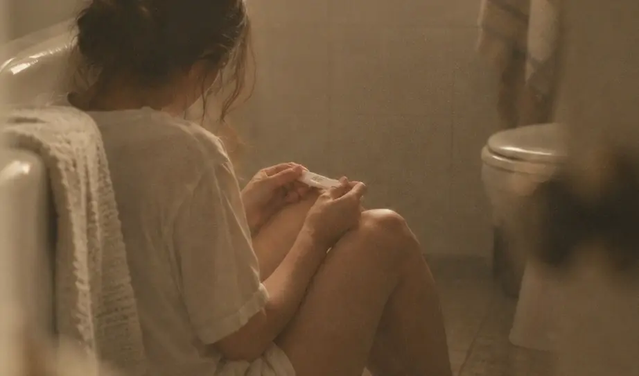 Mujer sentada en el suelo del baño, abrazando sus rodillas, con una prueba de embarazo en las manos